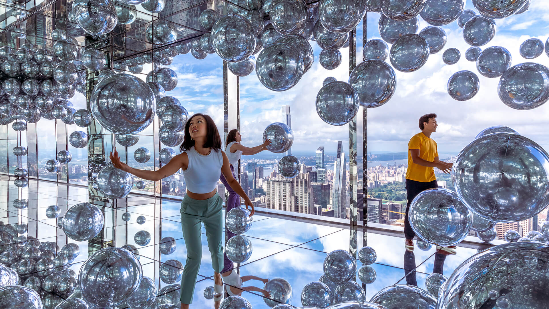 Visitors in the mirror room with silver balloons, reflecting themselves against the New York City skyline backdrop.