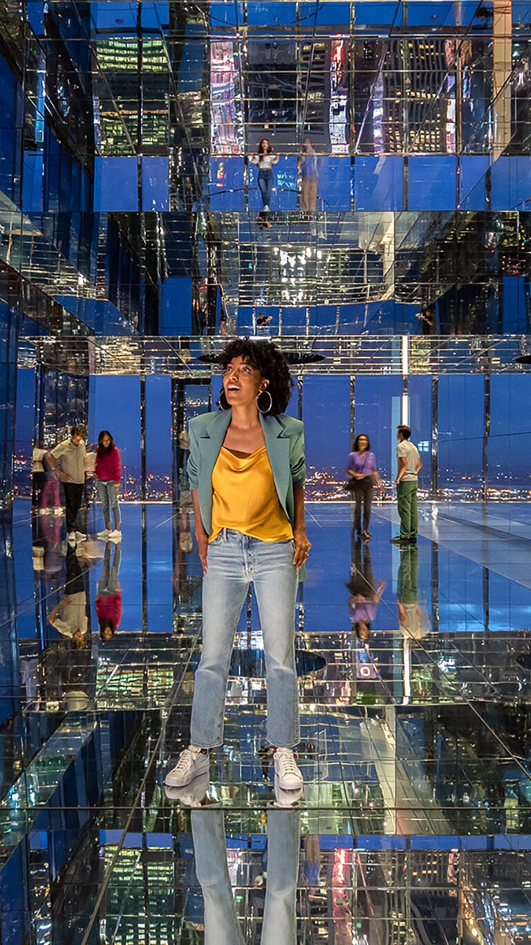 A woman marvels at the New York nightscape in a room of mirrors, where the city's lights multiply infinitely.
