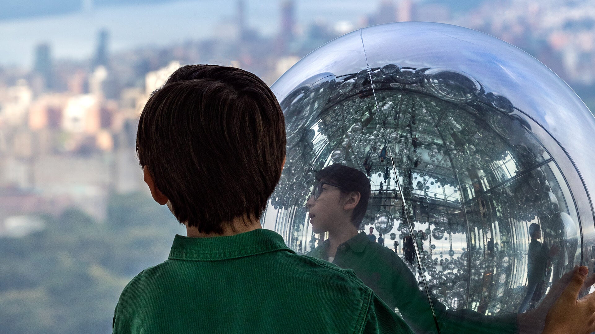 Young person in green shirt gazing into a large mirror ball, reflecting the New York City skyline in the background.