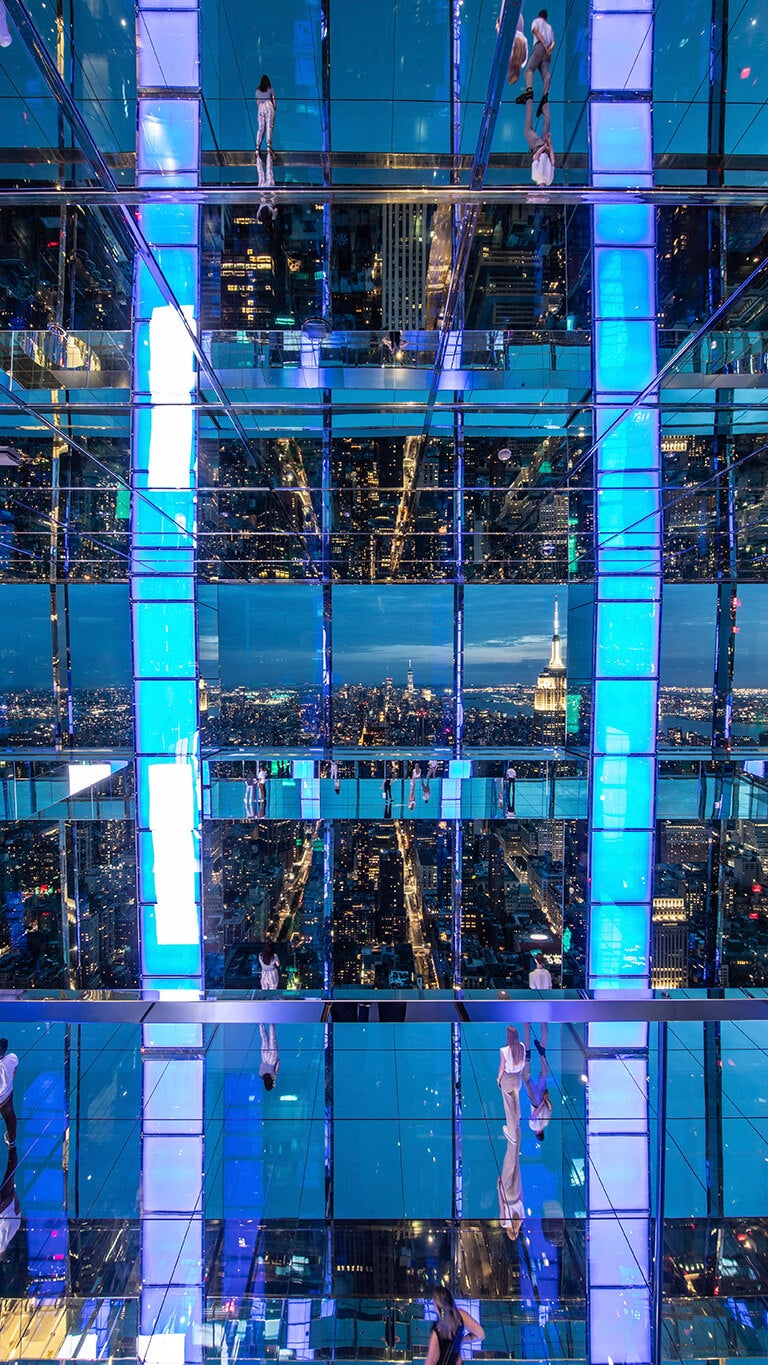 New York City night view from an observation deck with blue panels, showcasing the skyline and visitors.