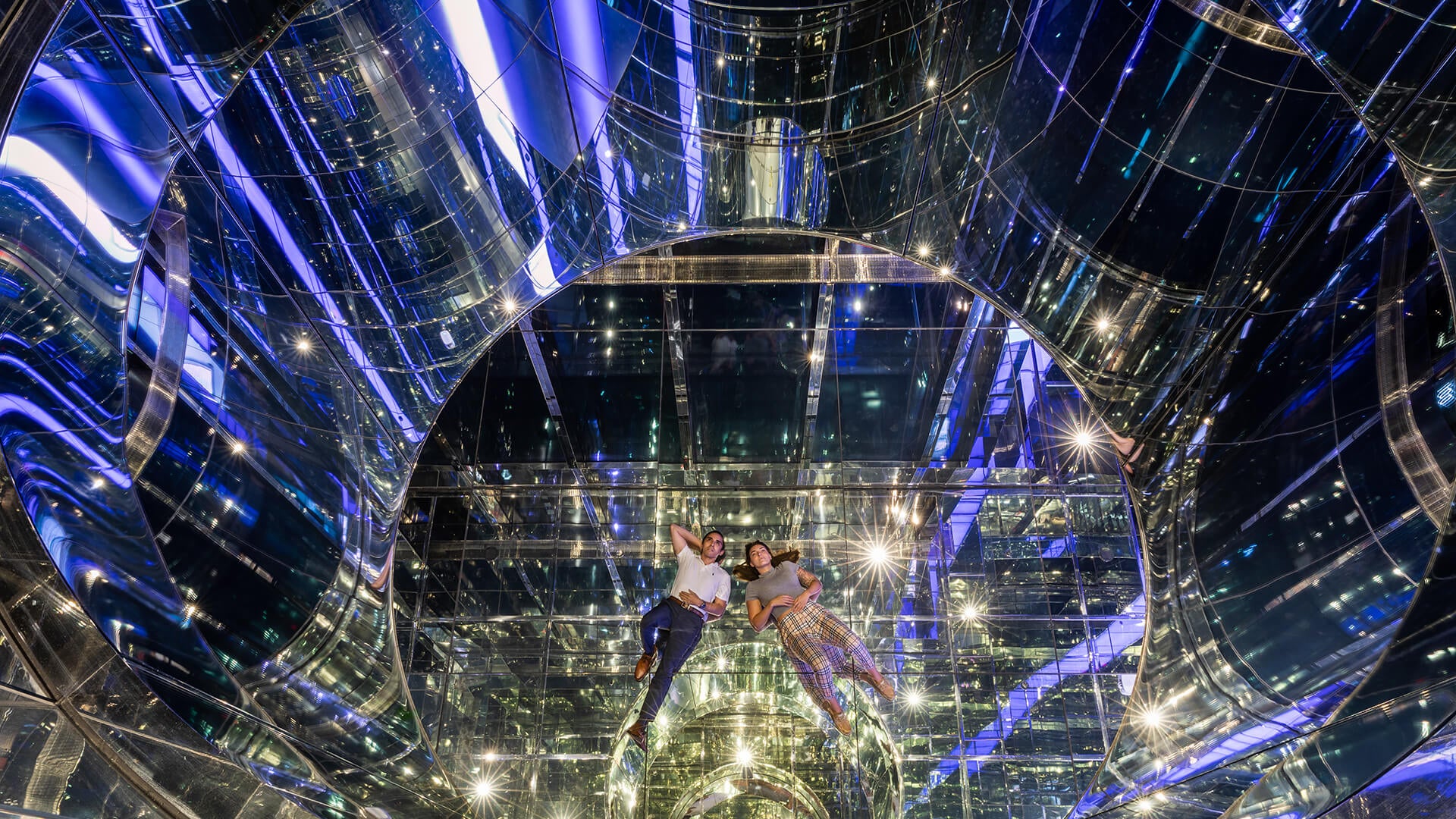 Two individuals floating in a reflective, transformative space illuminated by blue lights, showcasing art and architecture.