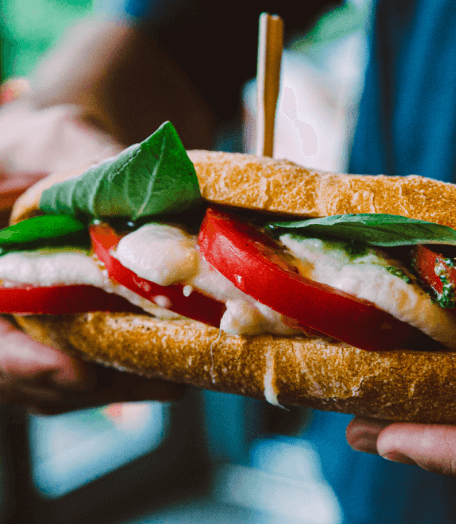 Close - up of a New York sandwich with fresh mozzarella, tomatoes, and basil, held in hand with a wooden skewer through it.