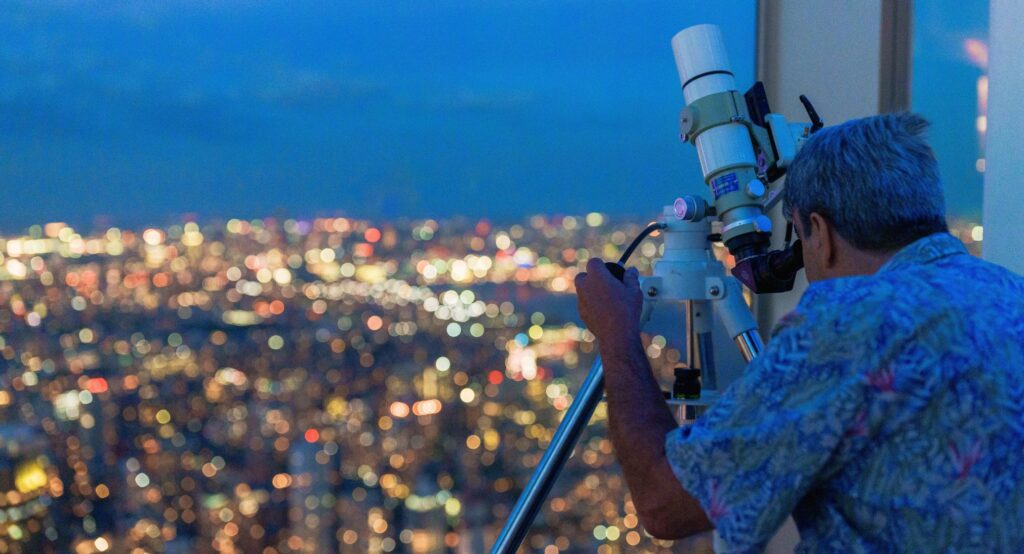 Man in floral shirt engaged in NYC stargazing with a telescope against the backdrop of a twinkling cityscape at dusk.
