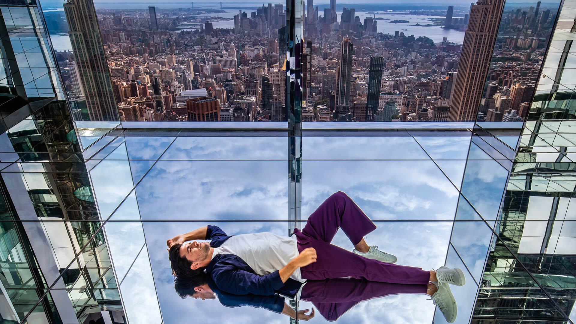 A man lounging on a transparent glass floor with the New York skyline reflecting in mirrored panels around him.