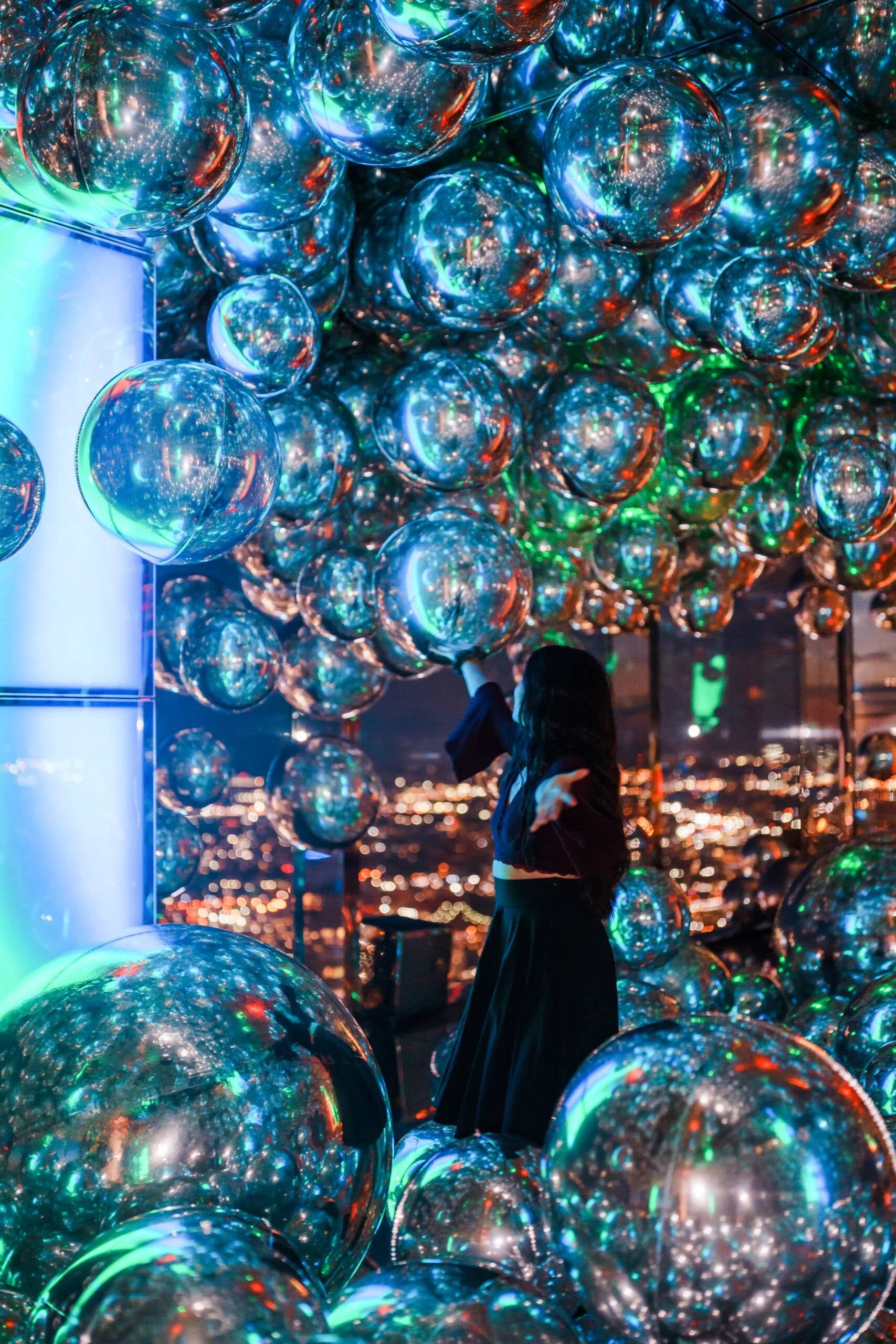 Person on an observation deck surrounded by reflective balloons, with city lights in the background.