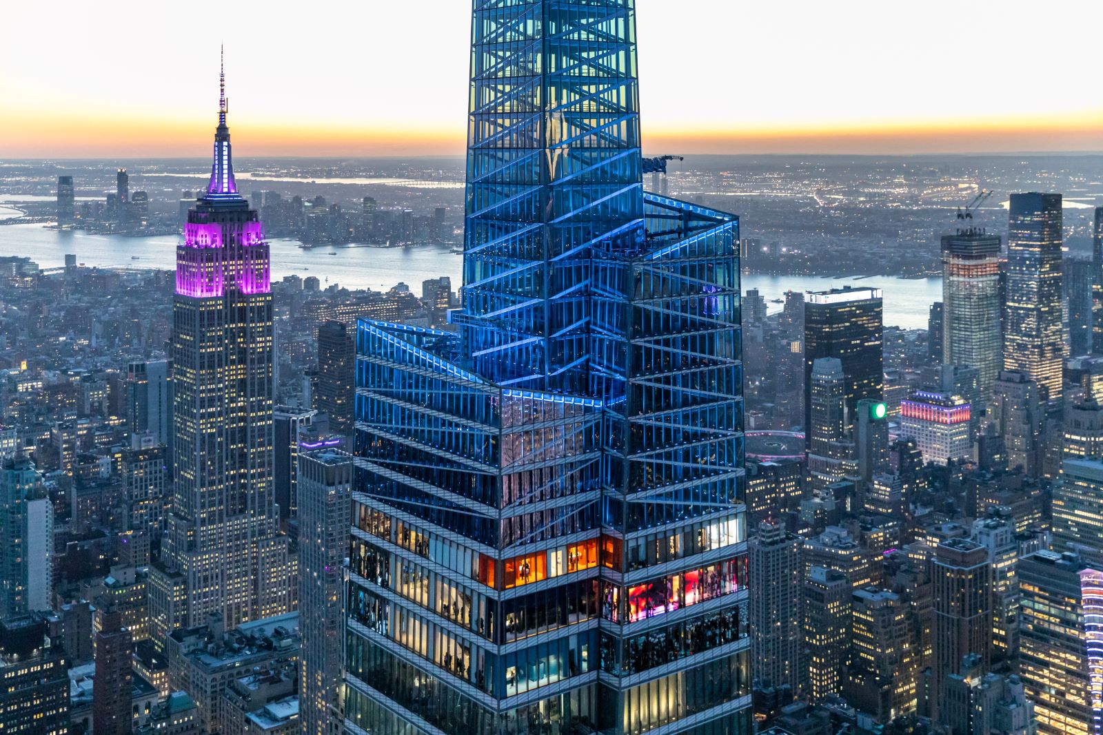 A view of the New York City skyline at sunset, featuring a brightly lit skyscraper and the Empire State Building.